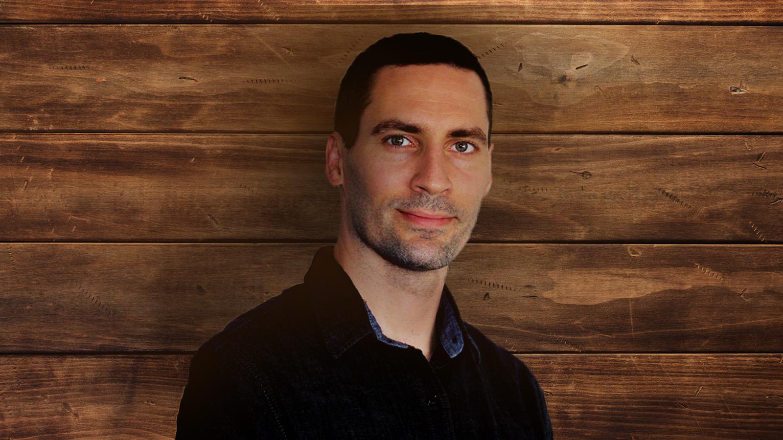 Justin Hosie standing in front of a paneled wood background. He is wearing a dark denim shirt and gently smiling.