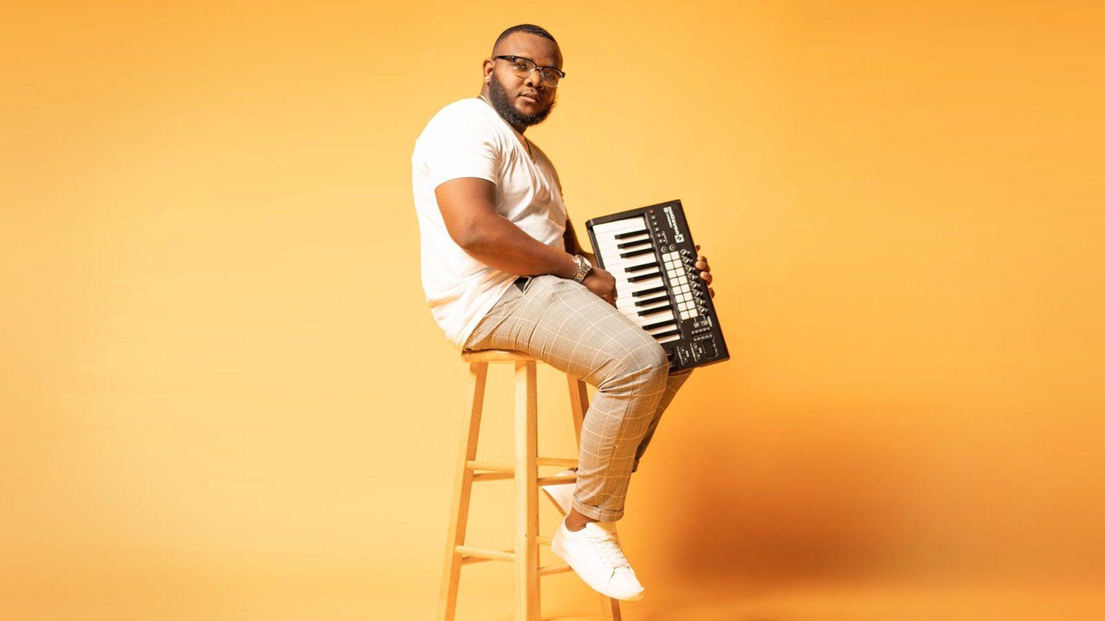 Chavez Parker sits on a stool and holds a keyboard. He wears glasses, a t-shirt, brown pants, and sneakers.