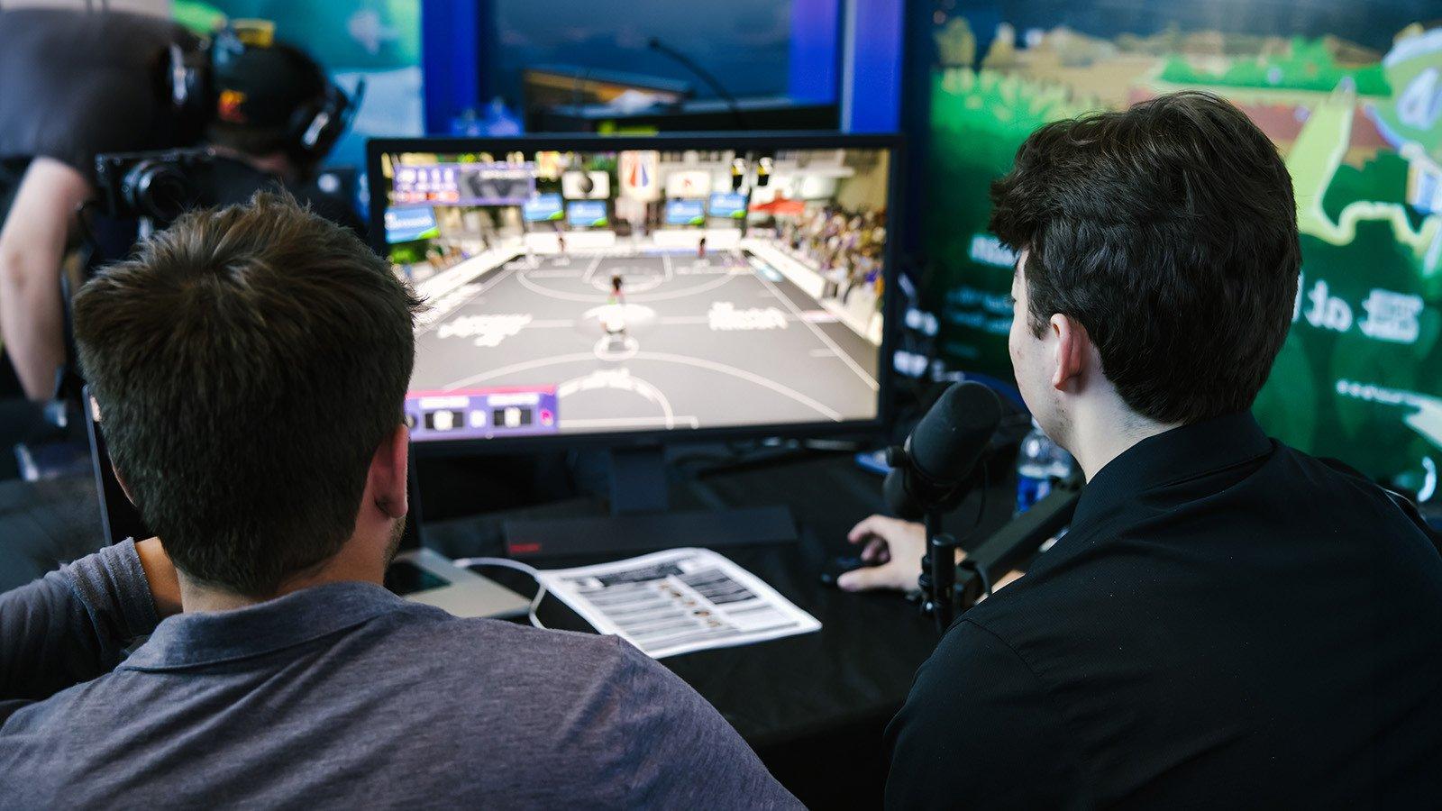 两个人背对着镜头，在他们面前桌子上的一个小显示器上观看“NBA 2 k”游戏.