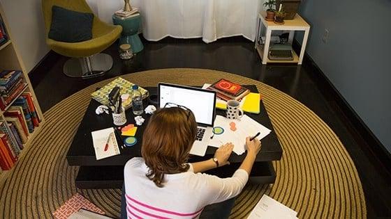 一个学生盘腿坐在咖啡桌前的地板上，手里拿着一台笔记本电脑, papers, notebooks, and pens.