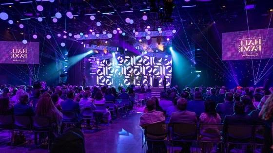 2023年名人堂入选者站在舞台上, surrounded by beautiful purple lighting in a large auditorium. 观众在鼓掌.