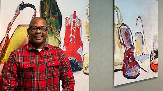 Anthony Roberts Jr. stands in front of two paintings of shoes. 他戴着一副黑框眼镜，穿着一件格子衬衫.