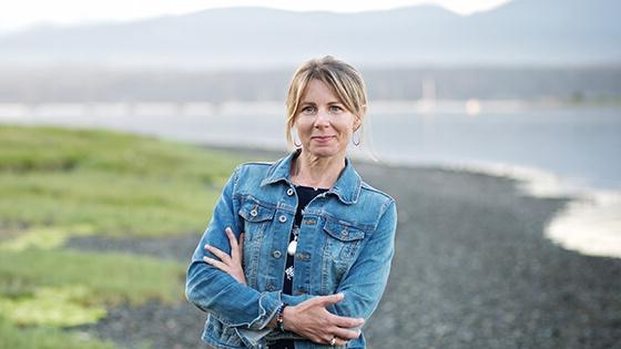 A blonde woman wearing a denim jacket over a navy top and black skirt stands with her arms crossed. She is outdoors in front of a river with mountains far in the background.