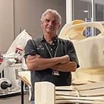 课程主任帕特·斯塔雷斯, 一个白发男子，身穿灰色polo衫, 站在一个大工作台旁边，在满帆的SimLab中，有一个f1赛车的3d打印复制品.
