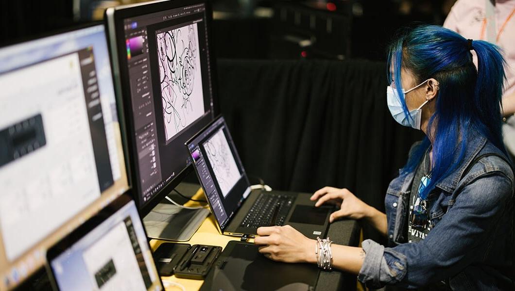 一个戴着口罩的女人, 一件t恤外搭一件牛仔夹克，放在笔记本电脑和台式显示器前. 屏幕上显示的是Adobe Photoshop中的图纸.